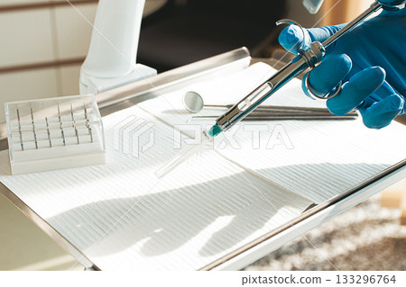The dentist's hand, in a blue glove, holds a carpal syringe on a tray, ready for a medical procedure. The dentist's hand, in a blue glove, holds a carpal syringe on a tray, ready for a medical procedure. 133296764
