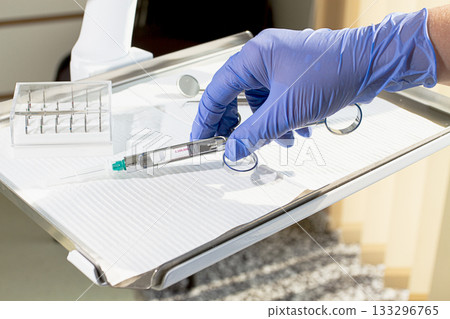 The dentist's hand, in a blue glove, holds a carpal syringe on a tray, ready for a medical procedure. The dentist's hand, in a blue glove, holds a carpal syringe on a tray, ready for a medical procedure. 133296765