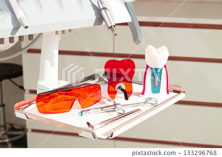On the tray are various dental instruments, a model dental implant and bright orange safety glasses, ready for use in clinical dental settings. On the tray are various dental instruments, a model dental implant and bright orange safety glasses, ready for use in clinical dental settings. 133296768