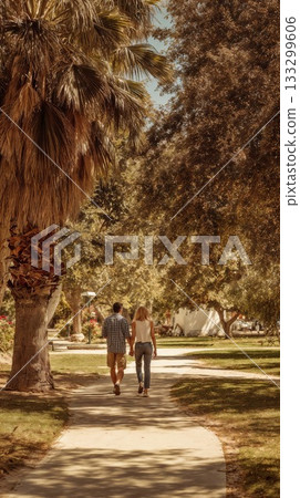 Young couple walking hand in hand through sunny park with palm trees and lush greenery, enjoying peaceful romantic outdoor date on warm sunny day together 133299606