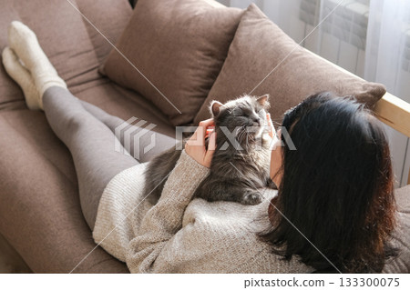 Woman is lying on brown sofa cuddling and stroking fluffy gray cat resting on her chest. Pet companionship, relaxation, cozy home life and comfort with animals Woman is lying on brown sofa cuddling and stroking fluffy gray cat resting on her chest. Pet companionship, relaxation, cozy home life and comfort with animals 133300075