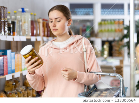 Girl customer in supermarket examines jar of pickled green olives, chooses food-stuff in grocery 133300564