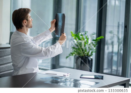 Male doctor sitting at desk looking at x-ray in modern clinic with window 133300635