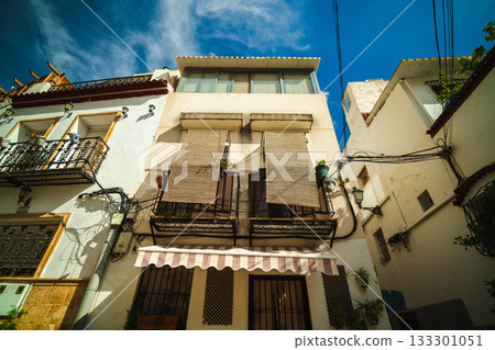 Residential facade in the Santa Cruz district of Alicante, Spain, with balconies, sunshades, and decorative plants against a bright blue sky. Residential facade in the Santa Cruz district of Alicante, Spain, with balconies, sunshades, and decorative plants against a bright blue sky. 133301051