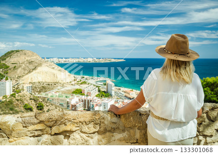 Woman wearing a hat enjoys the sea view and coastline from a scenic viewpoint in Alicante, Spain, on a bright sunny day. 133301068