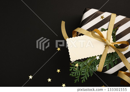 Fragment of Christmas black and white gift box with empty tag and cedar spruce on black background. Copy space Fragment of Christmas black and white gift box with empty tag and cedar spruce on black background. Copy space 133301555