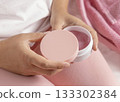 Closeup of woman in pink pyjamas holding cosmetic jar with blank lid in hands, mockup 133302384