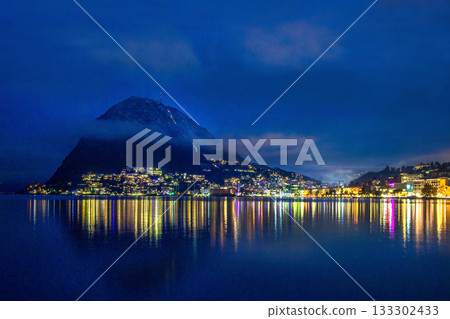 A stunning view of the Lugano waterfront and Monte San Salvatore during blue hour. Light fog. 133302433