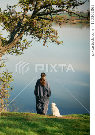 Back view of young woman and her small white dog relax and enjoy serene lake and autumn landscape 133302862