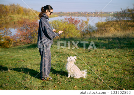Smiling woman training small white dog outdoors. 133302893