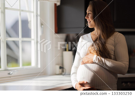 Smiling pregnant woman standing by a window in bright sunlight 133303250