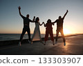 Four friends or family of four are jumping together at sunset near the ocean, holding hands and celebrating a joyful moment in silhouette. 133303907
