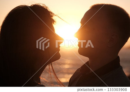 Silhouetted couple facing each other with the setting sun between them, creating a romantic and intimate golden hour moment. 133303908