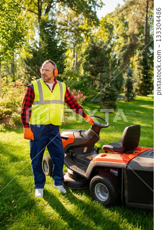 Worker in safety gear standing near lawn tractor in garden area 133304486