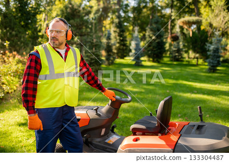 Male gardener standing near ride-on lawnmower on green grass 133304487
