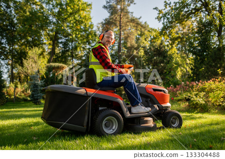 Worker in safety gear standing near lawn tractor in garden area 133304488