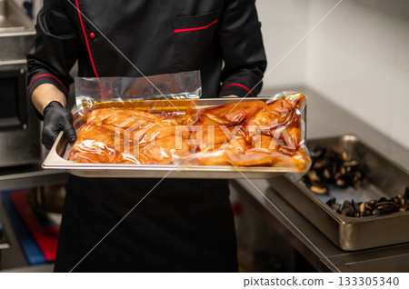 Chef in uniform holding metal tray with marinated food in kitchen Chef in uniform holding metal tray with marinated food in kitchen 133305340