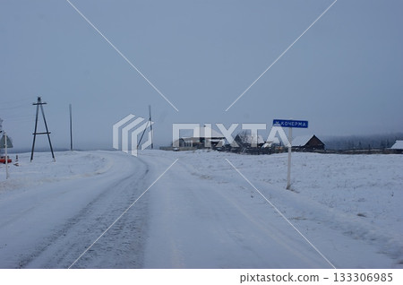 The photograph depicts old houses in a Siberian village, an old fence, and snowdrifts. The roofs of the houses are covered in a thick layer of snow. The photograph depicts old houses in a Siberian village, an old fence, and snowdrifts. The roofs of the houses are covered in a thick layer of snow. 133306985