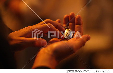 Close-up person hands gently holding a small, ornate locket against a warm background, evoking a sense of intimacy and mystery Close-up person hands gently holding a small, ornate locket against a warm background, evoking a sense of intimacy and mystery 133307659