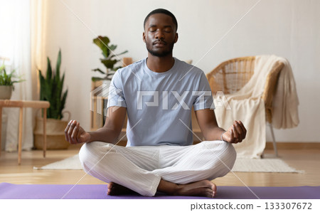 Mental health and self-care concept with a young man sits in lotus position on a yoga mat at home, meditating with eyes closed and holds his hands in Mudra. Mindfulness, inner calm and wellness 133307672