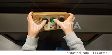 A child's hands grip a small wooden control panel with three buttons. Concepts include child development, tactile learning, technology for children, hand-eye coordination, and interactive exhibits. A child's hands grip a small wooden control panel with three buttons. Concepts include child development, tactile learning, technology for children, hand-eye coordination, and interactive exhibits. 133309440