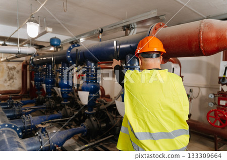 Engineer inspecting large blue industrial pipes and valves, safety vest and hard hat 133309664