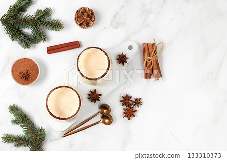 Eggnog with cinnamon, star anise, and nutmeg for Christmas and the winter holidays on a marble table with fir branches. A warming eggnog during the cold season, a festive arrangement for Christmas Eve Eggnog with cinnamon, star anise, and nutmeg for Christmas and the winter holidays on a marble table with fir branches. A warming eggnog during the cold season, a festive arrangement for Christmas Eve 133310373