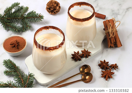 Eggnog with cinnamon, star anise, and nutmeg for Christmas and the winter holidays on a marble table with fir branches. A warming eggnog during the cold season, a festive arrangement for Christmas Eve 133310382