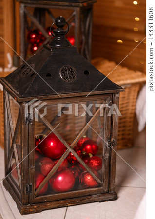 Vintage wooden lantern filled with red Christmas ornaments standing indoors as part of festive holiday d cor. Warm lights and wicker elements in the background create a cozy seasonal atmosphere. Vintage wooden lantern filled with red Christmas ornaments standing indoors as part of festive holiday d cor. Warm lights and wicker elements in the background create a cozy seasonal atmosphere. 133311608