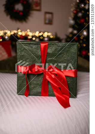A green wrapped gift box with a bright red ribbon sits on a white bedspread, surrounded by soft Christmas lights and festive holiday decor in the blurred background. A green wrapped gift box with a bright red ribbon sits on a white bedspread, surrounded by soft Christmas lights and festive holiday decor in the blurred background. 133311609