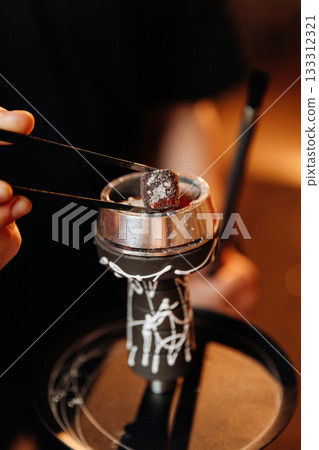 Hand placing hot hookah coal with tongs into metal bowl, warm bar bokeh light 133312321