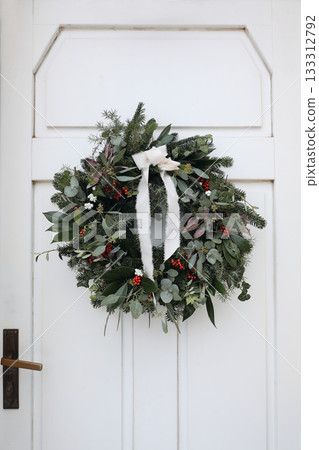 Lovely Christmas wreath. Rribbon, fir tree branches, eucalyptus, holly red berries hanging on white door. Floral still life. Natural festive decoration, december season, advent holiday. Vertical Lovely Christmas wreath. Rribbon, fir tree branches, eucalyptus, holly red berries hanging on white door. Floral still life. Natural festive decoration, december season, advent holiday. Vertical 133312792
