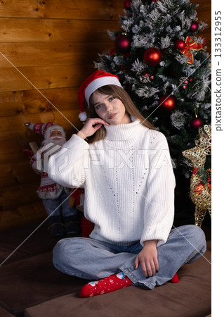 Little girl in a Santa hat seated near a Christmas tree 133312955