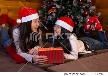 The two girls in Santa hats are joyfully exchanging a red gift box. The two girls in Santa hats are joyfully exchanging a red gift box. 133312966