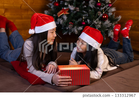 The two girls in Santa hats are joyfully exchanging a red gift box The two girls in Santa hats are joyfully exchanging a red gift box 133312967
