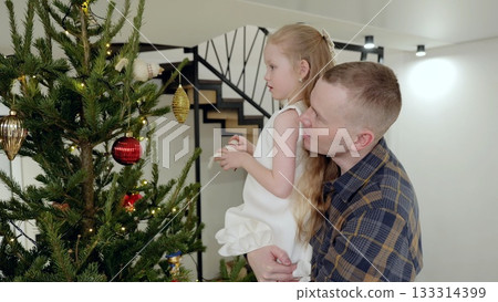 Parent supporting young child while hanging festive ornaments, sharing warm holiday bonding near decorated evergreen tree Parent supporting young child while hanging festive ornaments, sharing warm holiday bonding near decorated evergreen tree 133314399