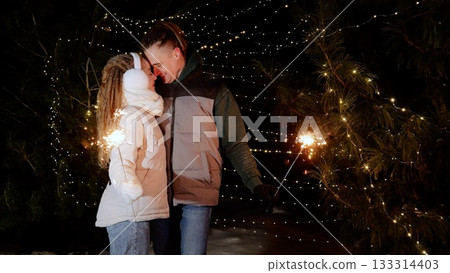 Couple holding sparkling fireworks, celebrating festive night amid illuminated winter forest with glowing holiday lights Couple holding sparkling fireworks, celebrating festive night amid illuminated winter forest with glowing holiday lights 133314403
