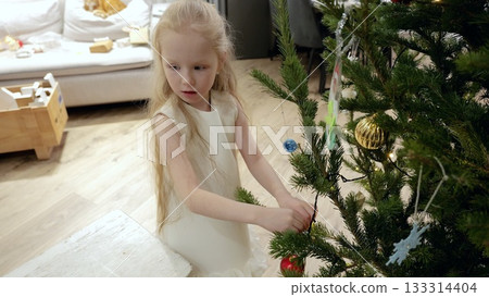 Young blond girl carefully hanging bright red ornament on decorated christmas tree, wearing white festive dress in warm, softly lit living room with holiday spirit Young blond girl carefully hanging bright red ornament on decorated christmas tree, wearing white festive dress in warm, softly lit living room with holiday spirit 133314404