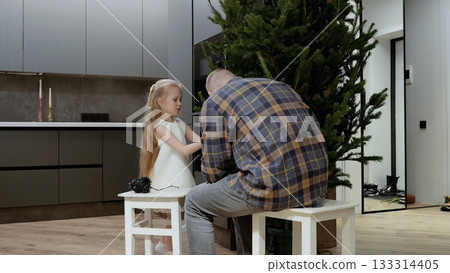 Father and daughter sitting on stools untangling christmas lights for the christmas tree in a modern apartment, preparing for the holiday season by decorating their home Father and daughter sitting on stools untangling christmas lights for the christmas tree in a modern apartment, preparing for the holiday season by decorating their home 133314405