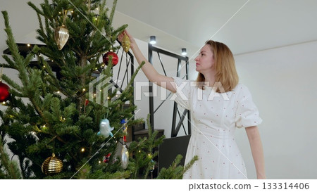 Woman wearing white dress hanging golden bauble on festively decorated christmas tree with twinkling lights, preparing holiday decor inside modern living space Woman wearing white dress hanging golden bauble on festively decorated christmas tree with twinkling lights, preparing holiday decor inside modern living space 133314406