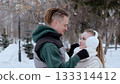Young man with dreadlocks adjusting the white scarf of his smiling girlfriend with earmuffs in a snowy park during winter, creating a romantic and cozy scene 133314412