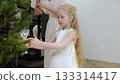Young girl with golden hair placing owl ornament on christmas tree, mother hanging decorations behind her in festive living room setting 133314417