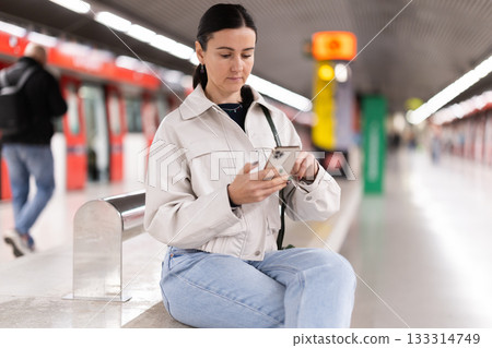 Girl with phone is sitting on metro platform station, scrolling though internet Girl with phone is sitting on metro platform station, scrolling though internet 133314749