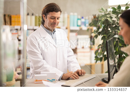 male pharmacist stands behind the counter near the computer and communicates with a customer 133314837