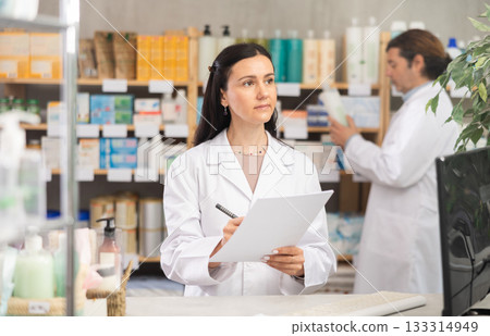Middle-aged female pharmacist restocking with paper and man working in background 133314949