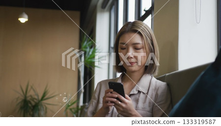 Sitting Asian woman browsing smartphone at lounge area, with potted plant and pendant light Sitting Asian woman browsing smartphone at lounge area, with potted plant and pendant light 133319587