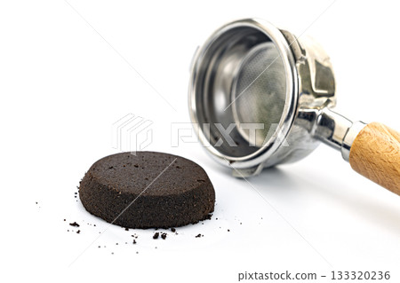 Used espresso coffee grounds puck alongside a stainless steel portafilter with wooden handle on white background 133320236