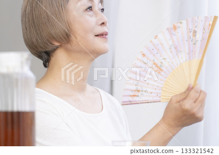 Senior woman cooling off with a fan in the living room 133321542