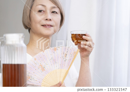 Senior woman cooling off with a fan in the living room Senior woman cooling off with a fan in the living room 133321547