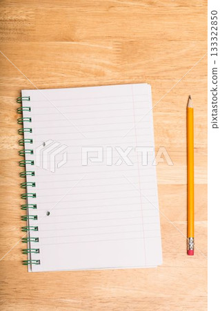 Spiral-bound lined notebook is lying open on wooden tabletop, with yellow pencil resting parallel 133322850